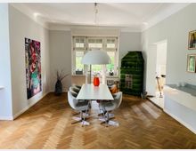 Helles Wohnzimmer mit Parkettboden, einem modernen Esstisch mit vier grauen Polsterstühlen, einer großen weißen Hängelampe und einem grün gekachelten Kamin. An der Wand hängt ein buntes Kunstwerk, durch ein großes Fenster fällt Tageslicht ein, zudem ist eine offene Verbindung zur Küche sichtbar.