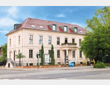 Historisches Gebäude mit heller Fassade namens "Palais Ritz", roten Ziegeldach und Dachgauben, umgeben von Bäumen und Zierpflanzen. Im Vordergrund befindet sich eine Straße mit Fußgängerüberwegen und einer Ampel.