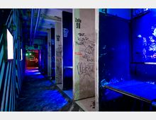 Ein schmaler, dunkler Gang in einem Gebäude, beleuchtet mit blauem Neonlicht. Die Wände und Betonpfeiler sind mit Graffiti bedeckt, einige Türen tragen die Aufschrift „Zelle“ mit Nummern; im Hintergrund ist ein Bereich mit weiteren bunten Lichtern erkennbar.