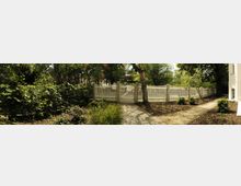 Ein grüner Gartenbereich mit einem weißen Holzzaun; ein gepflasterter Weg und ein mit Kies bestreuter Weg führen durch die Szene. Der Bereich ist von Bäumen und Sträuchern umgeben, angrenzend an ein helles Gebäude auf der rechten Seite.