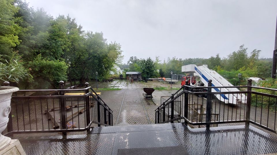 Eine Außenterrasse mit Metallgeländer und Treppe führt zu einem gepflasterten Hof, umgeben von Bäumen und Sträuchern. Im Hintergrund befinden sich ein kleines Gebäude, verschiedene Ausrüstungsgegenstände und eine Flugzeugtreppe, daneben steht ein roter Lkw.