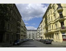 Straßenszene mit historischen Mietshäusern im Stil des 19. Jahrhunderts, links in dunklerem Beige und rechts in hellgelber Farbe mit Balkonen und Stuckverzierungen. Die Straße ist gepflastert und beidseitig von parkenden Autos gesäumt, im Hintergrund ein helles Gebäude vor einem bewölkten Himmel.