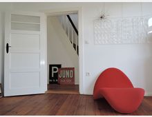 Helles Wohnzimmer mit Holzboden, einer roten, geschwungenen Designer-Sitzgelegenheit, einer weißen Tür mit Glaseinsatz und Blick auf eine Treppe im Hintergrund. An der Wand hängen moderne Kunstwerke, darunter eine weiße Skulptur und eine auffällige rote Retro-Werbetafel.