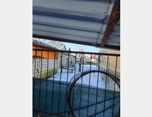 Ein verschneiter Hinterhof mit einem Metallzaun im Vordergrund, an dem ein Drahtbügel hängt. Im Hintergrund stehen kleine Gebäude aus weißem und orangefarbenem Mauerwerk, teils mit verschneiten Dächern.