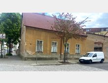 Eine alte, beige verputzte Hausfassade mit roten Dachziegeln und vier einfach verglasten Fenstern; davor ein gepflasterter Gehweg, ein kleiner Baum mit rosa Blüten und ein geparktes weißes Lieferfahrzeug. Im Hintergrund sind weitere Gebäude und eine gepflasterte Straße zu sehen.