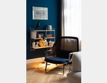 Ein modern eingerichtetes Zimmer mit blauer Wand, an der ein Regal mit Büchern, Dekorationsobjekten und einem Lautsprecher angebracht ist. Daneben steht ein Sessel mit schwarzem Stoffbezug auf einem hellen Parkettboden; im Hintergrund fällt Tageslicht durch einen weißen Vorhang.