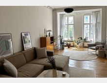 Helles Wohnzimmer mit Holzfußboden, einer beigen Eckcouch, einem runden Teppich und mehreren Zimmerpflanzen. Große Fenster mit weißen Vorhängen lassen viel Tageslicht hinein, und moderne Dekoration, darunter gerahmte Poster und Keramik, akzentuiert den Raum.