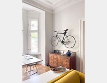 Bright, minimalist living room with high ceilings and ornate plaster molding, featuring tall white-framed windows and light wood floors. A wall-mounted bicycle hangs above a long wooden sideboard, with a white desk and chairs near the window and a sofa edge in the foreground.