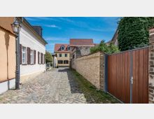 Schmale, gepflasterte Gasse in einer historischen Dorfumgebung mit dicht stehenden Häusern. Links befinden sich helle Gebäude mit Fenstern und Fensterläden, rechts eine Backsteinmauer und ein hölzernes Tor; im Hintergrund ein Haus mit roten Dachziegeln und mehreren Gauben.