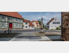 Eine Straßenszene in einem Dorf mit Fachwerkhäusern und roten Ziegeldächern. In der Mitte steht ein steinerner Obelisk auf einem gepflasterten Platz, umgeben von einer asphaltierten Straße.
