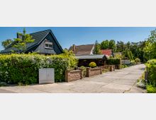 Straße mit Einfamilienhäusern, die durch niedrige Zäune und grüne Hecken voneinander getrennt sind; die Häuser haben Satteldächer in verschiedenen Farben und Gärten im Vordergrund, im Hintergrund sind Bäume und blauer Himmel sichtbar.