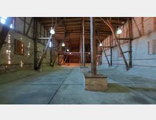 Ein großes, leerstehendes Gebäude mit einem Holzdachstuhl und freiliegenden Backsteinwänden. Mehrere Holzbalken und Säulen stützen die Konstruktion, während an der Decke Lampen hängen; der Boden ist aus Beton.