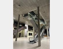 Großzügiger Innenraum eines modernen Gebäudes mit Sichtbetondecken, tragenden Säulen und einer Rolltreppe, die in das Obergeschoss führt. Der Boden ist mit hellen Fliesen bedeckt, und im Hintergrund befinden sich Wände mit Fenstern und Türen sowie ein Basketballkorb.