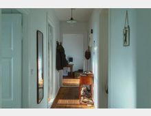 Ein heller Flur mit weißen Wänden und Holzboden; links hängt ein schmaler Wandspiegel, rechts sind ein kleiner Holztisch mit Gegenständen sowie Regale mit Schuhen sichtbar. Im Hintergrund führt der Flur zu einem weiteren Zimmer mit einem Holztisch.