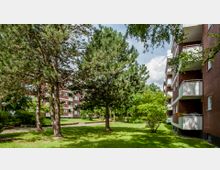 Mehrfamilienhäuser aus rotem Backstein mit Balkonen, umgeben von einer Grünfläche mit Bäumen, Sträuchern und gepflegtem Rasen; im Hintergrund ein wolkiger Himmel.