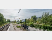Ein kleiner, unbedeckter Bahnhof namens "Yorckstraße" mit einem einzelnen Bahngleis, Sitzbänken und einem Hinweisschild. Im Hintergrund sind Bäume und vereinzelte Gebäude zu sehen, die von einer Grünfläche umgeben sind.