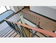 Treppe in einem mehrstöckigen Gebäude mit einem schwarz-geländerartigen Handlauf und einem orangeroten Holzgeländer. Der Bodenbelag besteht aus Linoleum und Fliesen, die Wände sind weiß gestrichen, und Tageslicht fällt durch große Fenster ein.