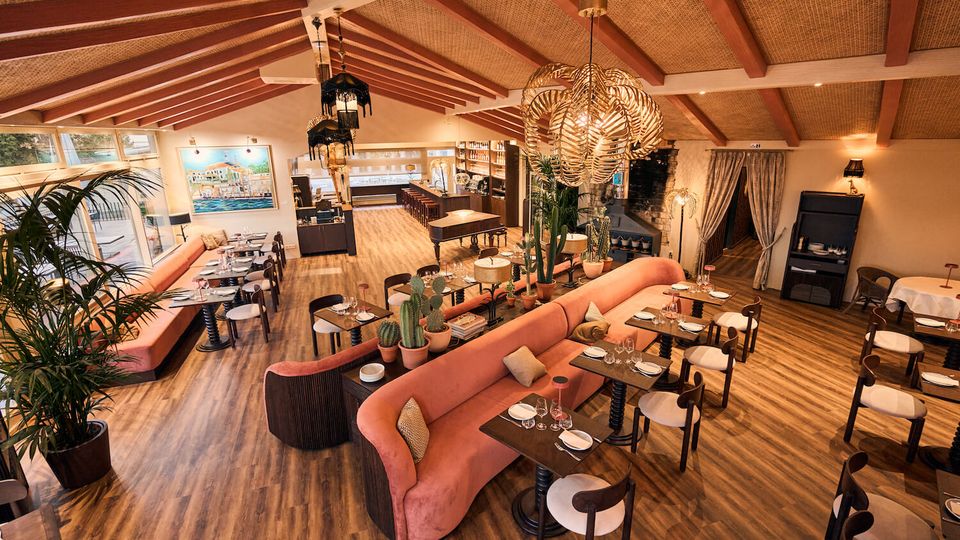 Spacious restaurant dining room with wood floors, exposed ceiling beams, and large woven pendant lights. Coral upholstered banquette seating surrounds set tables, with potted plants, tall windows along one side, and a bar area in the back.
