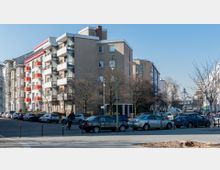 Straßenszene mit mehrstöckigen Wohngebäuden, teils mit modernen Balkonen und roten Akzentfarben. Im Vordergrund parkende Autos und ein Fahrradfahrer, im Hintergrund der Eingang einer U-Bahn-Station mit gewölbter Glaskonstruktion.