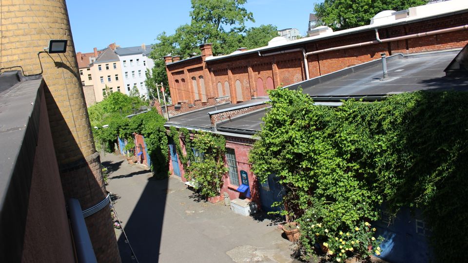 Innenhof eines ehemaligen Industrieareals mit niedrigen Backsteingebäuden und Flachdächern, teils mit Efeu und dichter Begrünung überwachsen. Links steht ein hoher runder Schornstein, im Hintergrund sind Wohnhäuser und große Bäume zu sehen.
