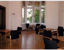 Heller Raum mit Parkettboden, mehreren runden Holztischen und schwarzen Stühlen mit Holzrahmen; große Fenster in einem Erker bieten Blick auf grüne Bäume draußen.