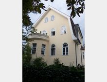 Gelbes zweistöckiges Wohnhaus mit Satteldach, großen weißen Rundbogenfenstern und einem kleinen Balkon mit Balustrade; im Vordergrund Bäume und dichter Gartenbewuchs.