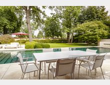 Moderner Gartenbereich mit einem rechteckigen Pool, einer Terrasse aus hellen Steinplatten und einem weißen Esstisch mit Stühlen im Vordergrund. Im Hintergrund befinden sich Bäume, Büsche und eine Rasenfläche, die von einer niedrigen Backsteinmauer begrenzt wird.