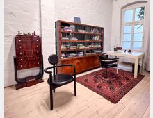 Ein minimalistisch eingerichteter Raum mit Holzboden, einem roten Orientteppich, einer Vitrine mit Büchern, einem glänzenden Holzschrank und einem weißen Schreibtisch neben einem großen Fenster. Zwei moderne Stühle, darunter ein schwarzer Sessel, stehen im Raum.