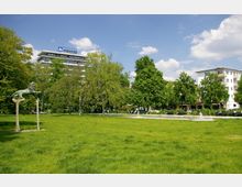 Grüne Parklandschaft mit Rasenfläche im Vordergrund, einer Löwenskulptur aus Metall links und Bäumen im Hintergrund. Hinter den Bäumen sind ein modernes Bürogebäude mit „Hochtief“-Logo sowie Wohnhäuser sichtbar.