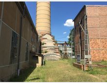 Industriegebäude mit Backstein- und verputzten Fassaden, flankiert von einem hohen, runden Schornstein aus gelben Ziegeln. Im Vordergrund eine Wiese mit vereinzelten Metallstrukturen und Rohren, die auf eine industrielle Nutzung hinweisen.