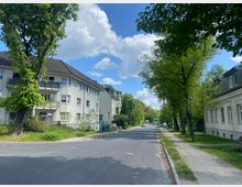 Straßenansicht in einem Wohngebiet mit mehrstöckigen Wohnhäusern auf der linken und rechten Seite, umgeben von Bäumen. Der asphaltierte Gehweg und die Rasenflächen entlang der Straße ergänzen die ruhige, grüne Umgebung unter einem bewölkten blauen Himmel.