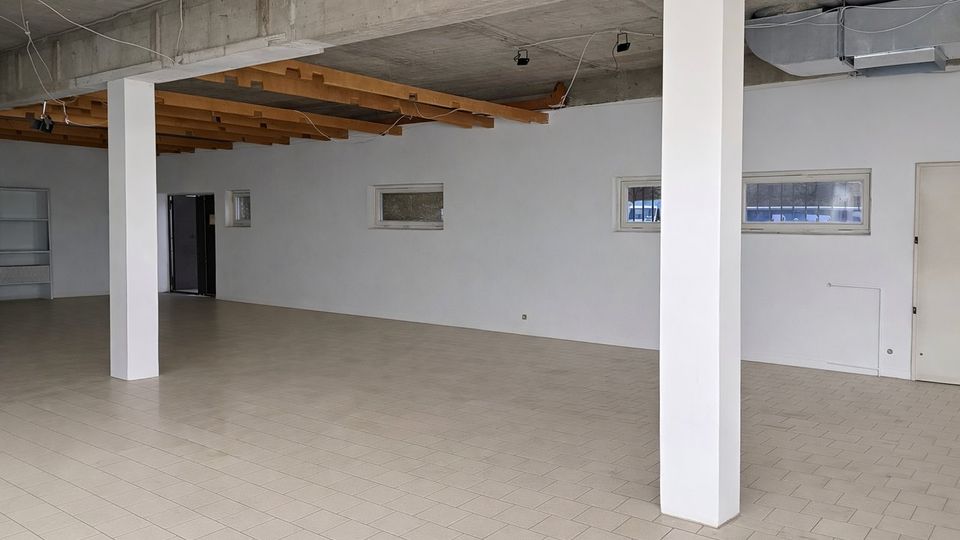 Großer, leerer Innenraum mit hellem Fliesenboden, weißen Wänden und mehreren weißen Stützsäulen. Sichtbare Betondecke mit freiliegenden Holzbalken, Kabeln und Lüftungskanal sowie schmale horizontale Fenster und Türen an der Rückwand.