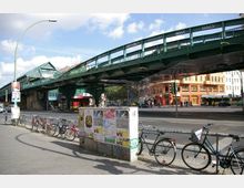 Ein erhöhtes Bahngleis aus grünem Metall dominiert die Szene einer städtischen Kreuzung, mit einer darunterliegenden Straße und Gehwegen. Im Vordergrund sind geparkte Fahrräder sowie eine Plakatwand, im Hintergrund bunte Gebäude und vereinzelt Passanten sichtbar.
