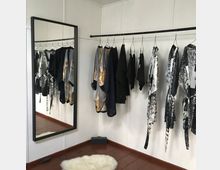 Ein minimalistisch eingerichteter Raum mit weißer Wand und Holzfußboden, in dem auf einer schwarzen Stange Kleidung in neutralen und gemusterten Farben hängt. Ein großer rechteckiger Spiegel lehnt an der Wand, und ein flauschiger, weißer Teppich liegt auf dem Boden.