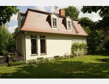 Einfamilienhaus mit hellgelber Fassade und rotem Ziegeldach. Vor dem Haus befindet sich ein gepflegter Garten mit Rasen, Sträuchern und Kletterpflanzen, umgeben von Bäumen.