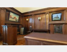 Innenraum eines historischen Saals mit holzvertäfelten Wänden, einem Rednerpult im Vordergrund und mehreren Gemälden an den Wänden. Im Raum befinden sich außerdem eine Holzbank, ein Stuhl und Wandlampen mit klassischem Design.