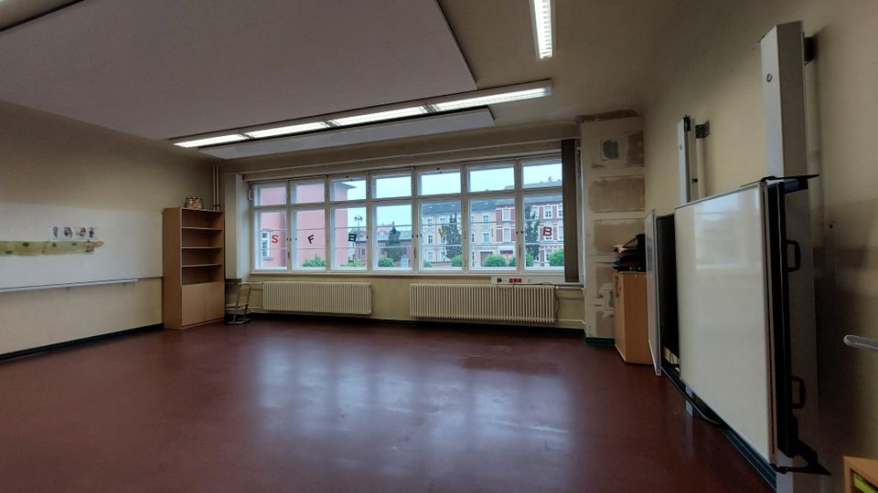 Ein leerer Klassenraum mit rotbraunem Boden, großen Fenstern an einer Wand, mehreren Heizkörpern darunter und Blick auf Gebäude außerhalb. An der rechten Seite befinden sich ein Whiteboard und ein Schrank, links an der Wand hängt eine leere Tafel.