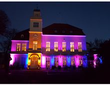 Beleuchtetes historisches Gebäude mit cremefarbener Fassade und rotem Dach im Dunkeln, angestrahlt von bunten Lichtern in Violett, Blau und Gelb. Das Gebäude hat große, rechteckige Fenster, einen zentralen Eingang mit Rundbogen sowie einen kleinen Turm mit einer Wetterfahne an der Spitze.