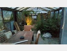 Innenraum eines Gewächshauses mit einem Holztisch und Stühlen im Vordergrund. Links und rechts sind Pflanzen und Gartenutensilien zu sehen, der Bereich ist schwach beleuchtet.