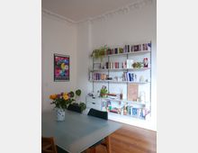 Wohnzimmer oder Essbereich mit einem großen Glastisch und vier Stühlen, dekoriert mit einem Blumenstrauß in einer Vase. Im Hintergrund befindet sich ein Wandregal mit Büchern, Pflanzen und Dekorationsgegenständen; die weiße Wand hat eine kunstvolle Stuckleiste an der Decke.