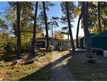 Ein Gartenbereich mit hohen Kiefern, einer kleinen weißen Hütte mit großen Fenstern im Hintergrund sowie einem grünen, zeltähnlichen Gebäude rechts. Im Vordergrund befinden sich Sitzbänke, eine Schaukel und eine Holzbank.