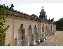Barocke Fassade eines Gebäudes mit hellgelbem Putz, verzierten Fenstern, Reihen von Skulpturen und Vasen auf Marmorsockeln. Auf dem Dach befinden sich Zierelemente und eine Uhr in einem kupfergedeckten Turm, umgeben von Grünflächen.