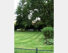 Ein gepflegter Park mit einer großen Rasenfläche, umgeben von hohen Bäumen und Sträuchern. Im Hintergrund stehen weiße Sitzbänke vor dicht bewachsenen Heckenbögen.