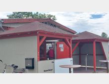 Ein kleiner Imbissstand mit rot-weißen Holzpavillons und einer offenen Verkaufsfläche. Im Vordergrund stehen runde Stehtische, daneben ist ein Fahrrad teils sichtbar.