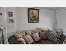 Wohnzimmerbereich mit dunklem Sofa, vielen Kissen und Decken vor einer weißen Wand. Darüber hängen ein großes gerahmtes Landschaftsbild und mehrere kleinere gerahmte Fotos; links steht ein Glasregal, rechts ein kleines Beistelltischchen mit Lampe.