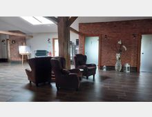 Ein gemütlicher Innenraum mit roten Backsteinwänden, dunklem Holzfußboden und sichtbaren Holzbalken. Im Vordergrund stehen Sessel um einen kleinen Tisch, im Hintergrund befinden sich eine Staffelei, eine Sitzbank, eine Glastür und Deko-Elemente wie ein großer weißer Blumenstrauß in einer Vase.