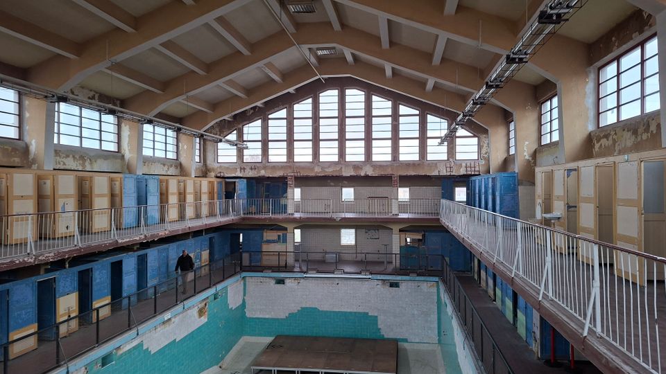 Innenansicht eines alten, leeren Schwimmbads mit bröckelnden Wänden, einem leeren Becken und einer umlaufenden Galerie mit Geländern. Große Fensterreihen lassen Tageslicht in den Raum, während Umkleidekabinen mit abgenutzten Türen die Wände säumen.