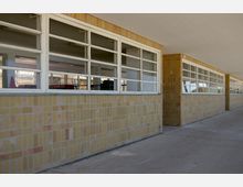 Langer Gebäudeflur mit gelben Klinkerwänden und großen rechteckigen Fenstern mit weißen Rahmen, die in einen Innenraum mit sichtbaren Möbeln blicken. Der Boden besteht aus glattem Beton, und die Decke ist schlicht gehalten.