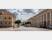 Straßenszene in einer Altstadt mit Kopfsteinpflaster, gesäumt von historischen Gebäuden mit pastellfarbenen Fassaden und Mansarddächern. Kleine Baumreihen und schmiedeeiserne Laternen prägen das ruhige Straßbild, unter einem leicht bewölkten Himmel.