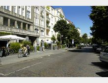 Straßenszene in einer Wohngegend mit gepflasterter Fahrbahn, flankiert von historischen Gebäuden mit Balkonen auf der linken Seite und Bäumen auf der rechten. Im Vordergrund sind Fahrräder, ein Café mit Sonnenschirmen und Passanten zu sehen.